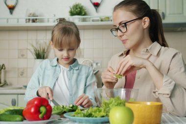 Cute loving daughter is helping mother with salad cutting cucumbers feeding young woman and laughing having fun at home. People and cookery concept.